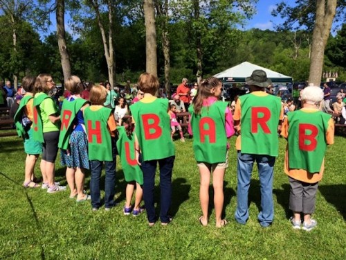 A group of people in green shirts with red letters spelling "RHUBARB" stands together outdoors at a community event.