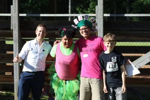 Four people pose together outdoors, smiling and wearing colorful outfits, with a festive atmosphere in the background.
