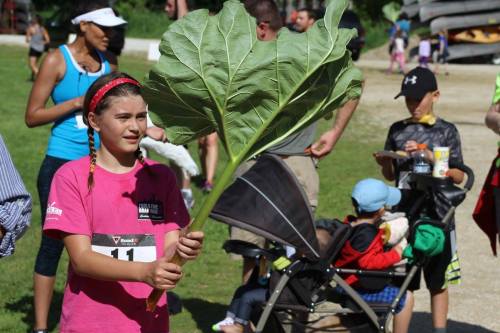 A girl in a pink shirt holds a large leaf while people gather in the background at an outdoor event.