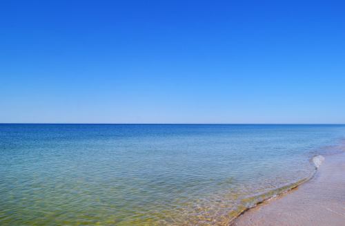 A serene beach scene with clear blue water and a cloudless sky, gently lapping waves on the sandy shore.