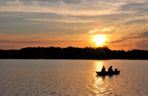 Silhouetted fishermen in a boat on a calm lake at sunset, with vibrant colors reflecting on the water.