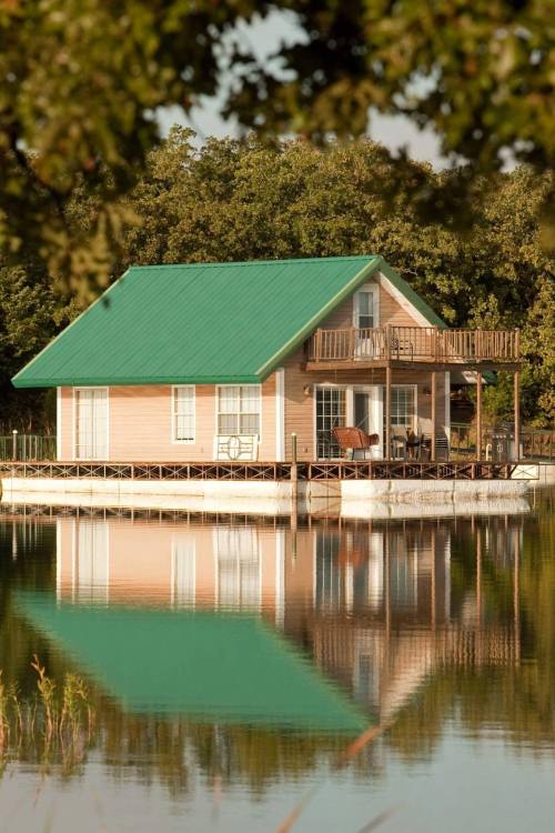 A charming lakeside cabin with a green roof, surrounded by trees, reflecting in the calm water.