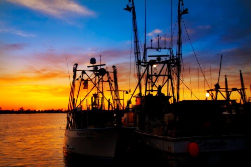 Fishing boats docked at sunset, silhouetted against a colorful sky with vibrant orange and blue hues.