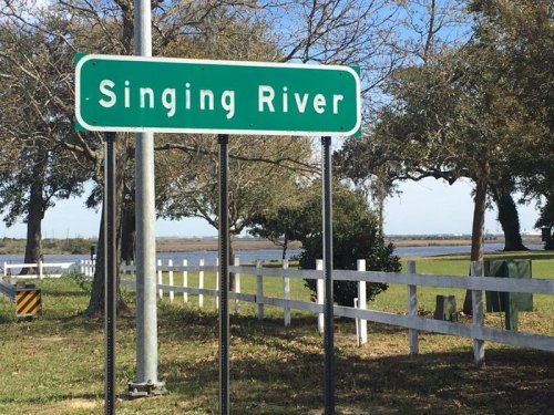 A green road sign reading "Singing River" near a white fence and trees, with a river visible in the background.