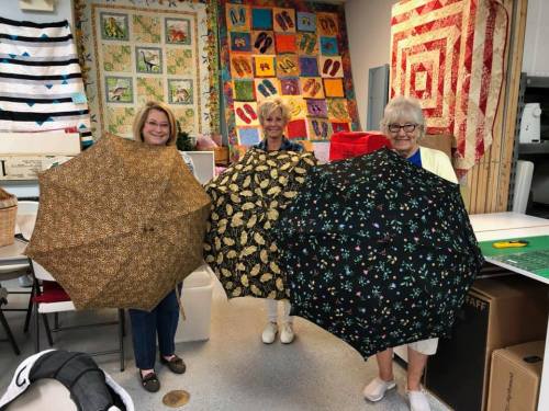 Three women hold colorful umbrellas in a craft room filled with quilts and fabric.