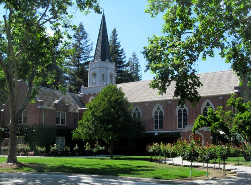A brick building with a tall spire, surrounded by trees and a garden, under a clear blue sky.