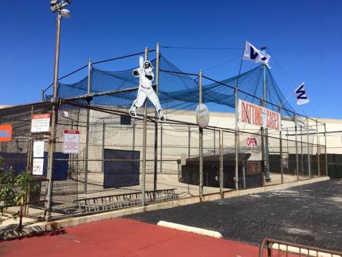 A batting cage facility with a blue sky, featuring a baseball player silhouette and flags, marked "Open."