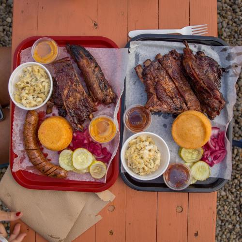 Two trays of barbecue featuring ribs, sausage, cornbread, macaroni, pickles, and sauces on a wooden table.