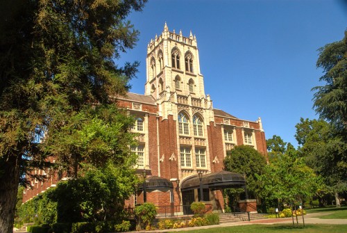 Historic brick building with a tall clock tower, surrounded by trees and blue sky.