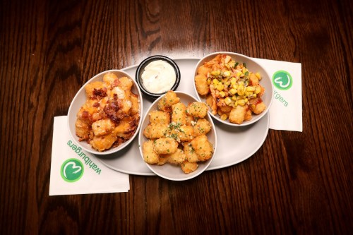 Three bowls of tater tots with various toppings, served with a dipping sauce on a wooden table.