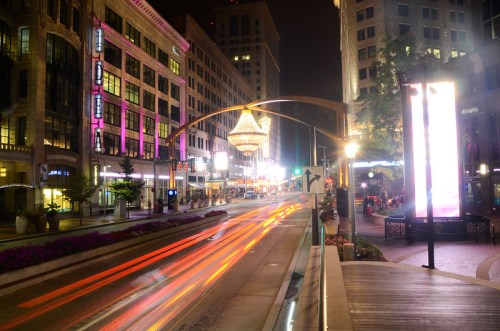 A vibrant city street at night, featuring colorful lights, a large chandelier, and streaks of car headlights.