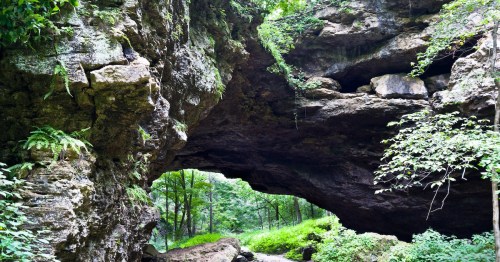 A natural rock arch surrounded by lush greenery and trees in a serene forest setting.