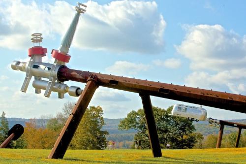 A large metal sculpture resembling machinery, with a cable car in the background against a scenic landscape.