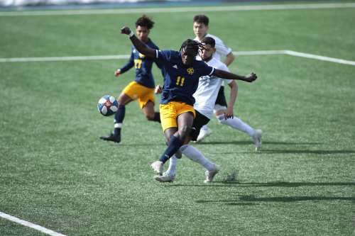 A soccer player in a blue jersey kicks the ball while two opponents challenge him on a green field.