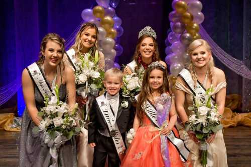 A group of six pageant winners, including a young boy and girl, holding flowers and wearing sashes, smiling together.