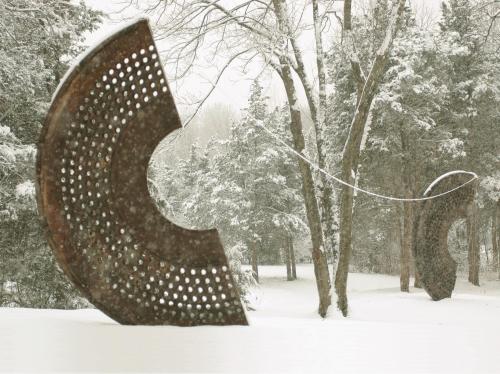 Two large, curved metal sculptures in a snowy landscape, surrounded by trees and falling snowflakes.