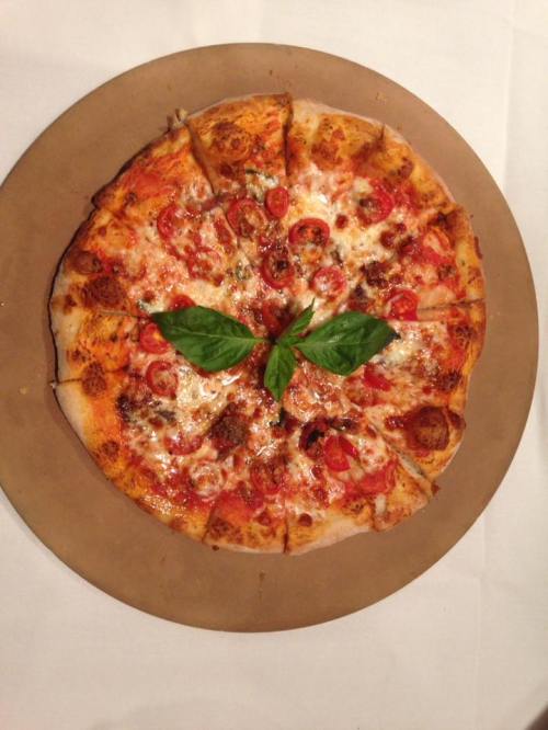 A round pizza topped with cheese, tomatoes, and fresh basil, served on a brown paper plate.