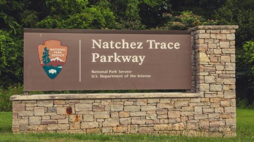 Sign for Natchez Trace Parkway, featuring the National Park Service logo and stone structure, surrounded by greenery.