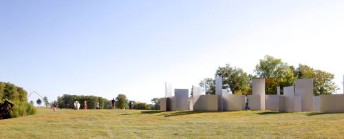 Visitors explore a large outdoor sculpture installation on a grassy field under a clear blue sky.