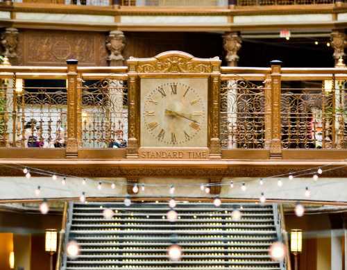 A decorative clock with Roman numerals, labeled "STANDARD TIME," hangs above a staircase in an ornate building.