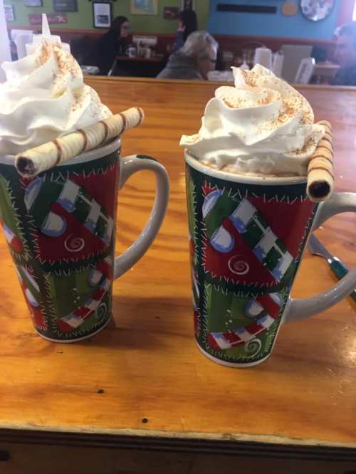 Two festive mugs filled with whipped cream and topped with a rolled wafer, sitting on a wooden table.