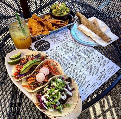 A plate of colorful tacos with toppings, tortilla chips, guacamole, and a drink on a table with a menu.