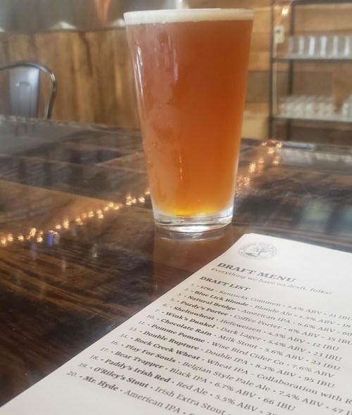A pint of amber beer sits on a wooden table next to a draft menu featuring various beer options.