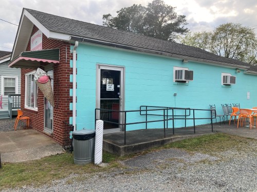 A turquoise building with a brick facade, featuring a ramp, air conditioning units, and orange chairs outside.