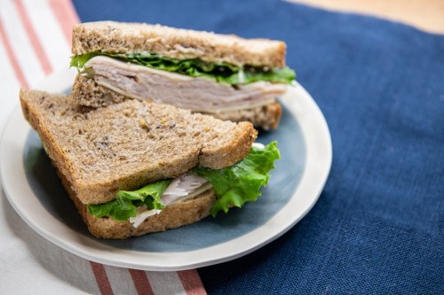 A plate with a sandwich made of whole grain bread, turkey, and lettuce, resting on a blue tablecloth.