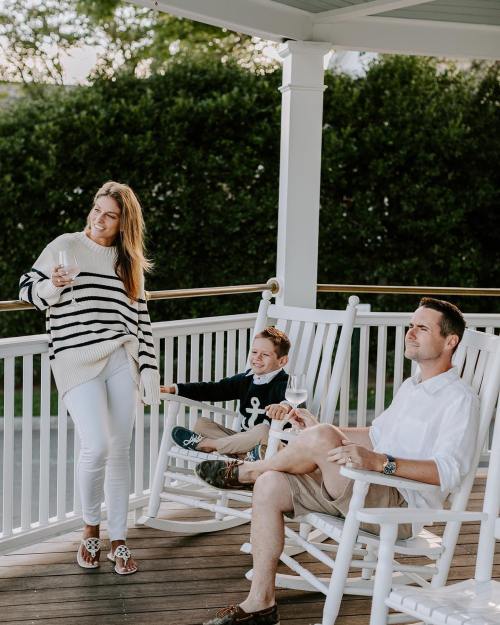 A woman in a striped sweater and white pants stands with a glass, while a man sits in a rocking chair with a child.