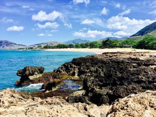 Scenic beach view with rocky shoreline, clear blue water, and lush green hills under a partly cloudy sky.