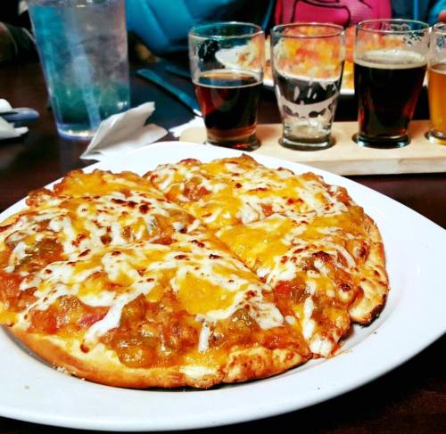 A close-up of a cheesy pizza with toppings, served on a white plate, with a flight of craft beers in the background.