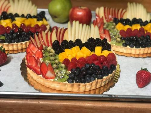 Colorful fruit tarts topped with strawberries, blueberries, kiwi, mango, and apple slices on a baking tray.