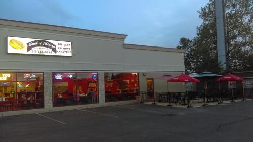 Exterior of a restaurant with a sign, outdoor seating, and a parking lot at dusk.