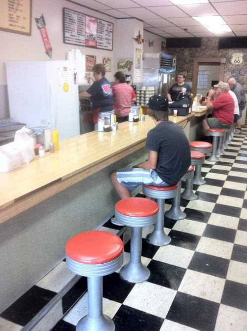 A retro diner with a long counter, red stools, and patrons enjoying food and drinks. A person sits alone at the counter.
