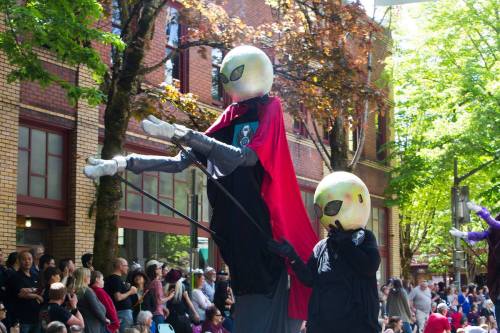 Two performers in oversized alien masks and capes, walking on stilts during a lively outdoor parade.