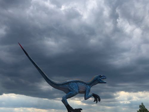 A blue dinosaur model with a long tail stands against a backdrop of dark, cloudy skies.
