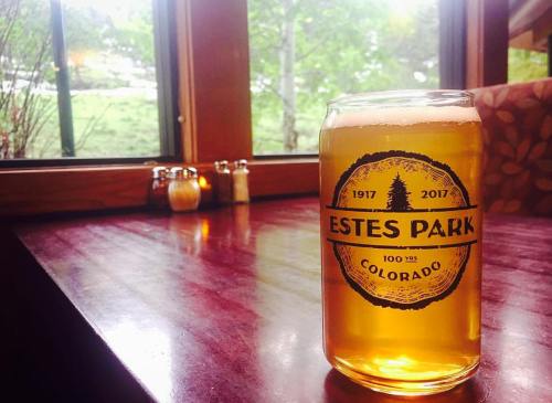 A glass of beer sits on a wooden table with a view of greenery outside a window, featuring an "Estes Park" label.