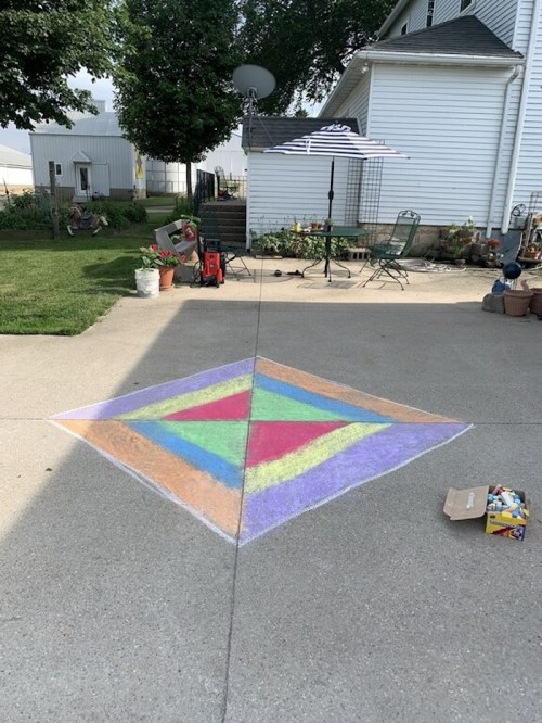 Colorful chalk drawing of a diamond shape on a concrete driveway, with an umbrella and garden furniture in the background.