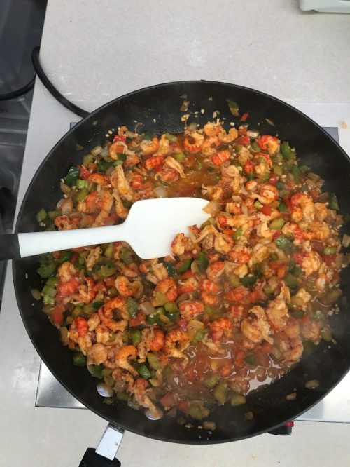 A skillet filled with shrimp and colorful diced vegetables, cooking on a stovetop with a white spatula.