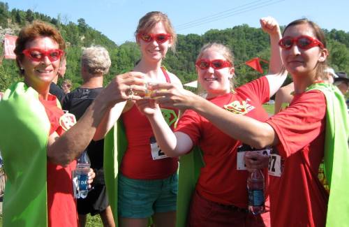Four women in red shirts and green capes celebrate with drinks, wearing sunglasses, at an outdoor event.