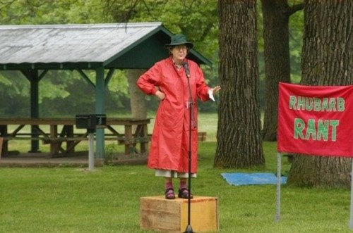 A person in a red raincoat stands on a wooden box, speaking into a microphone at a park event titled "Rhubarb Rant."