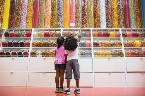 Two children reach for colorful candy in a vibrant candy store filled with jars and containers of sweets.