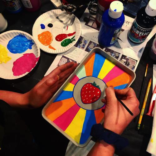 A person paints a colorful strawberry design on a tray, surrounded by paint palettes and art supplies.