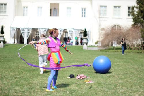 A performer in colorful attire twirls ribbons on a grassy lawn, with a large blue ball nearby and people in the background.