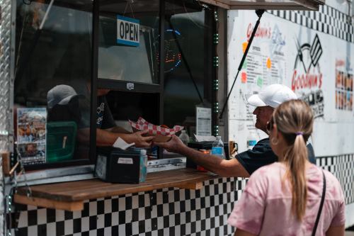 A customer receives food from a food truck window, with a sign reading "OPEN" and a checkered pattern in the background.
