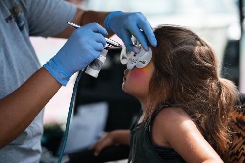A child receives face painting with an airbrush, while an artist carefully applies colorful designs.