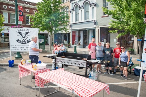 A group of people grilling at a street fair, with a banner for Haren's Market in the background and tables set up.