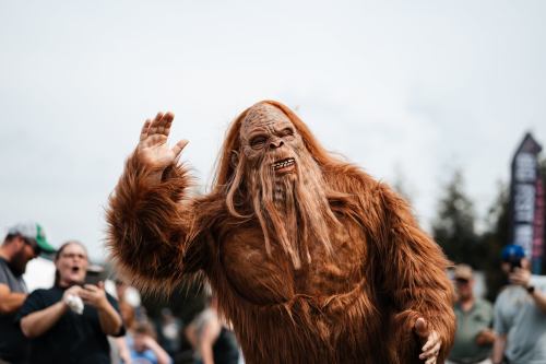 A person in a Bigfoot costume waves to a crowd at an outdoor event, surrounded by onlookers.