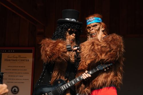 Two costumed figures, one in a top hat with a guitar and the other in a headband, pose playfully at a heritage center.